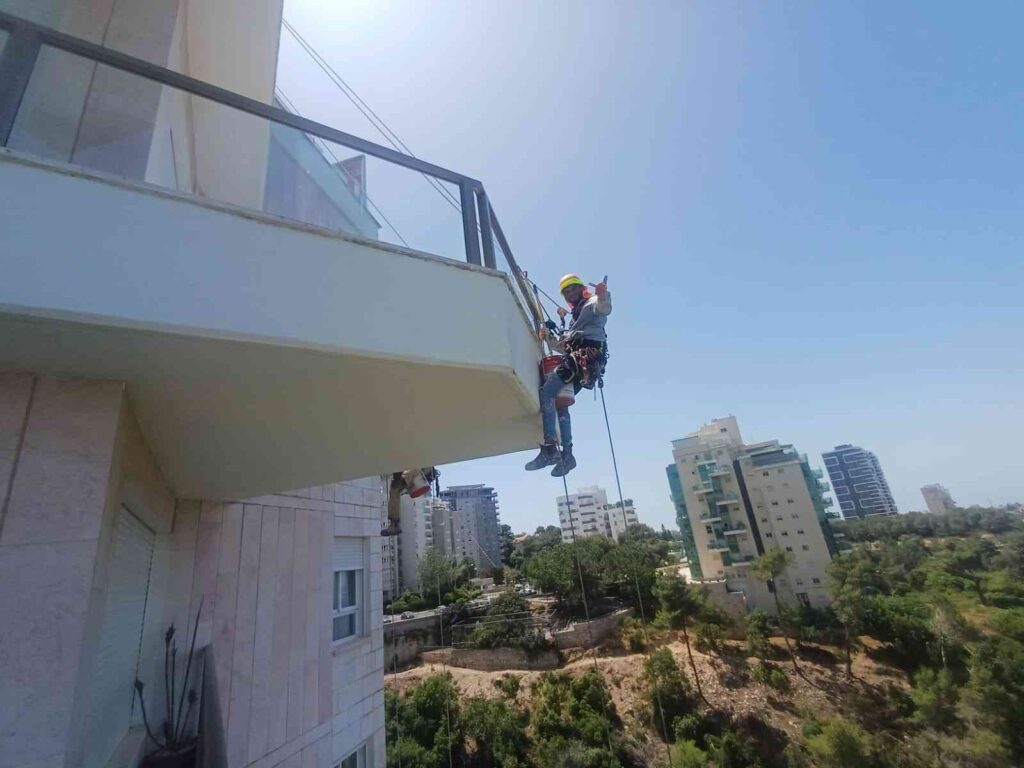עובד מבצע עבודות סנפלינג בגובה על קיר חיצוני של בניין באזור מגורים.