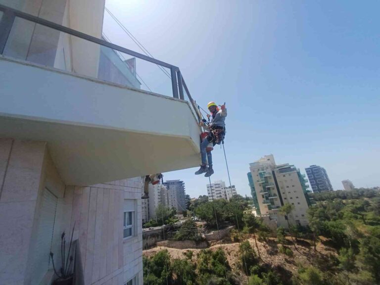 עובד מבצע עבודות סנפלינג בגובה על קיר חיצוני של בניין באזור מגורים.